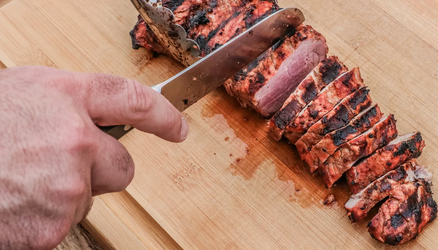 Mão fatiando carne grelhada com faca sobre tábua de madeira